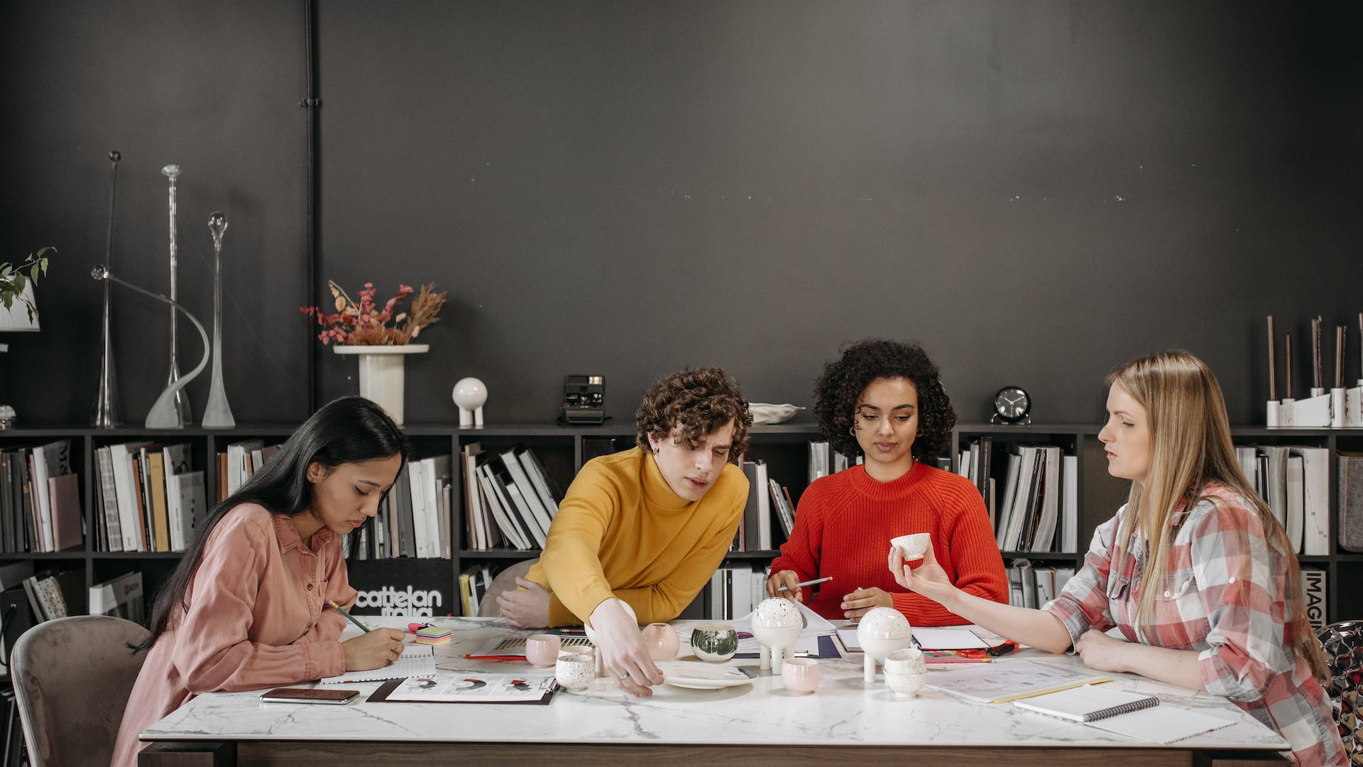 Four creative professionals engaged in a collaborative meeting around a table, surrounded by books and design materials, brainstorming ideas to harness the concept of 'Herd Mentality' in their project.