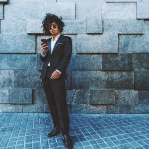 An entrepreneur in a chic black suit and sunglasses stands casually against a textured stone wall, smartphone in hand, representing the epitome of Mastering Visual Content for Dynamic Social Media.