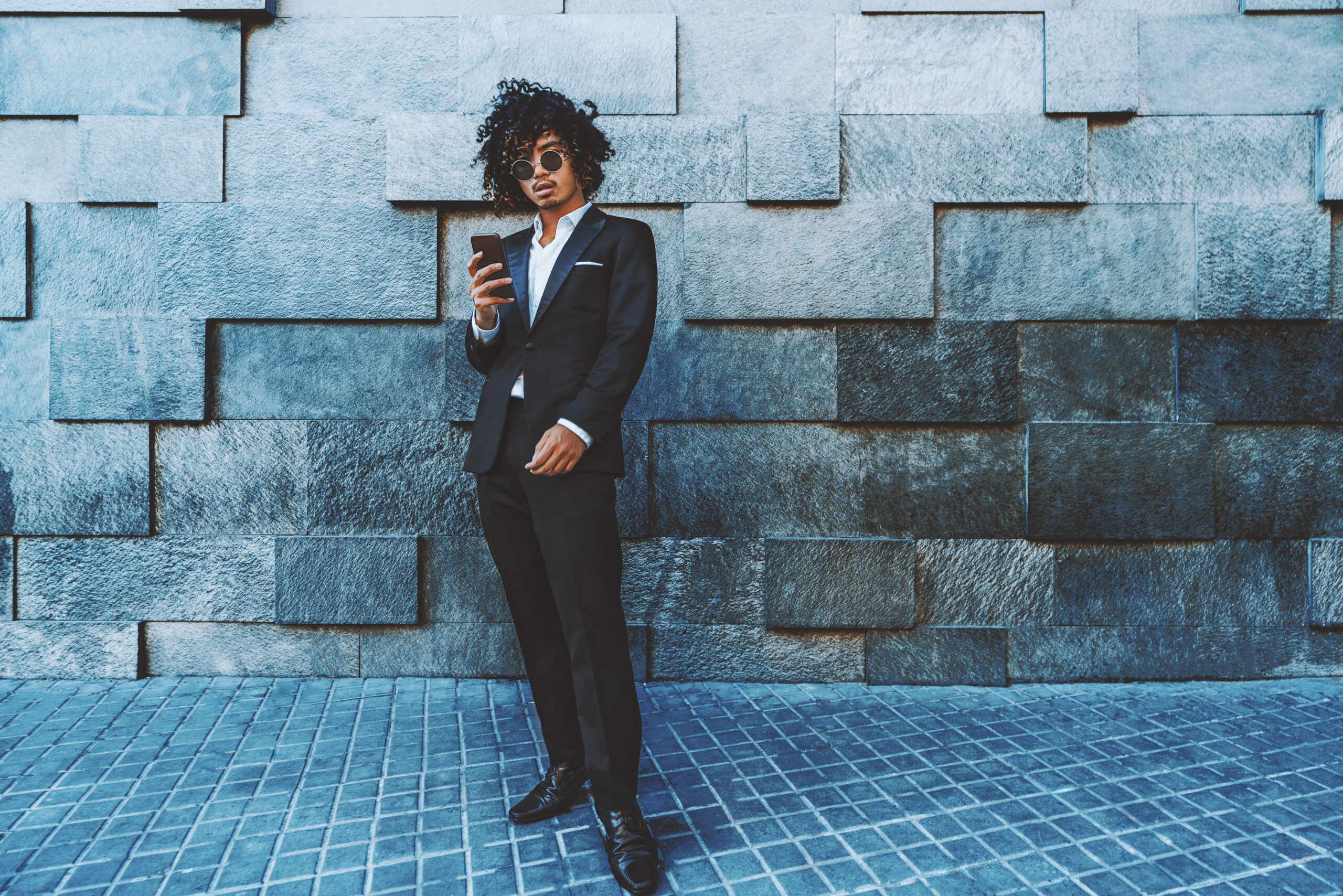 An entrepreneur in a chic black suit and sunglasses stands casually against a textured stone wall, smartphone in hand, representing the epitome of Mastering Visual Content for Dynamic Social Media.