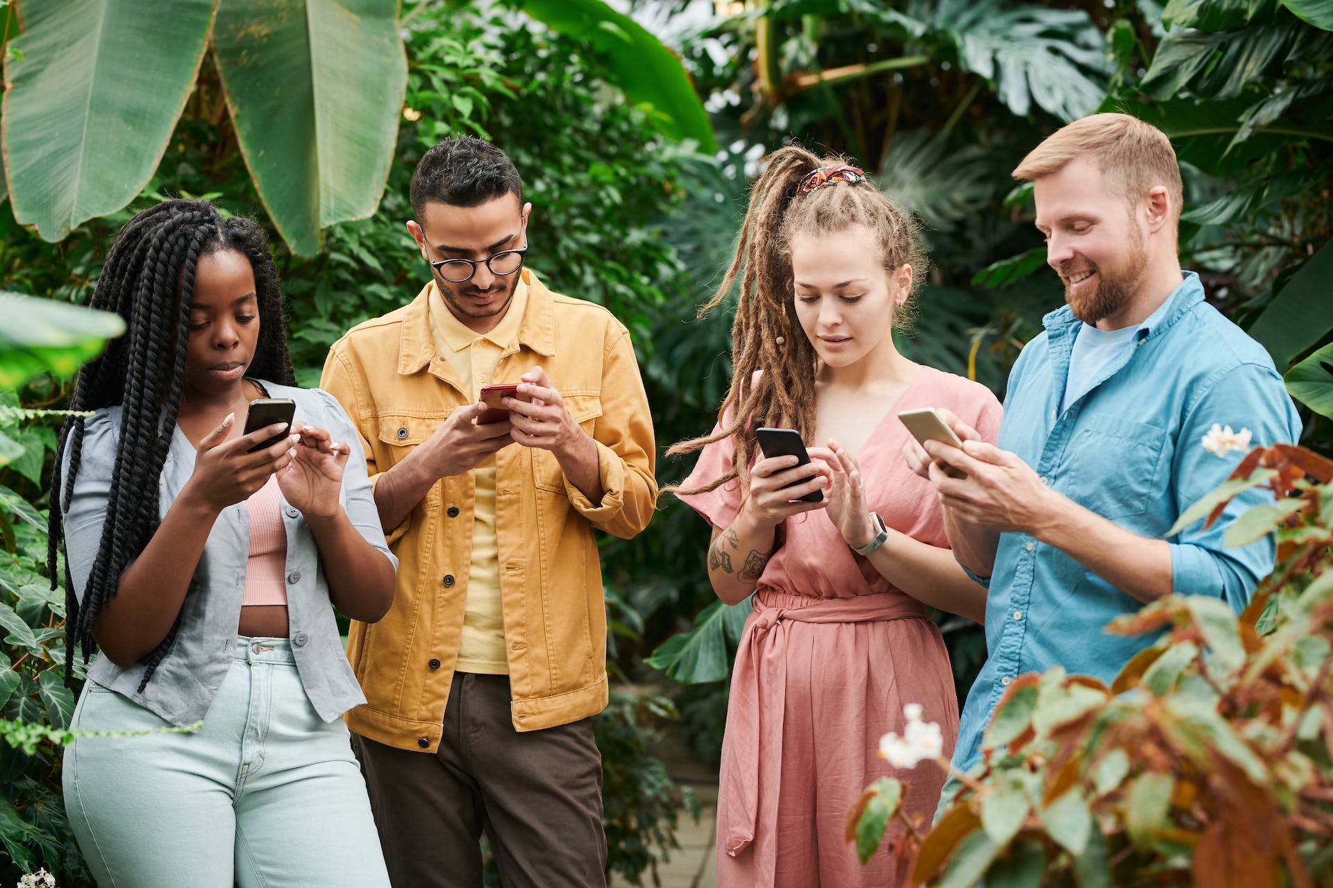 A diverse group of four people in a lush greenhouse, each focused intently on their smartphones. They stand amid vibrant green foliage, suggesting a relaxed yet engaging atmosphere of learning and connectivity. This image evokes a sense of community and the shared pursuit of knowledge, embodying the collaborative spirit of the "Instagram Branding Seminar".