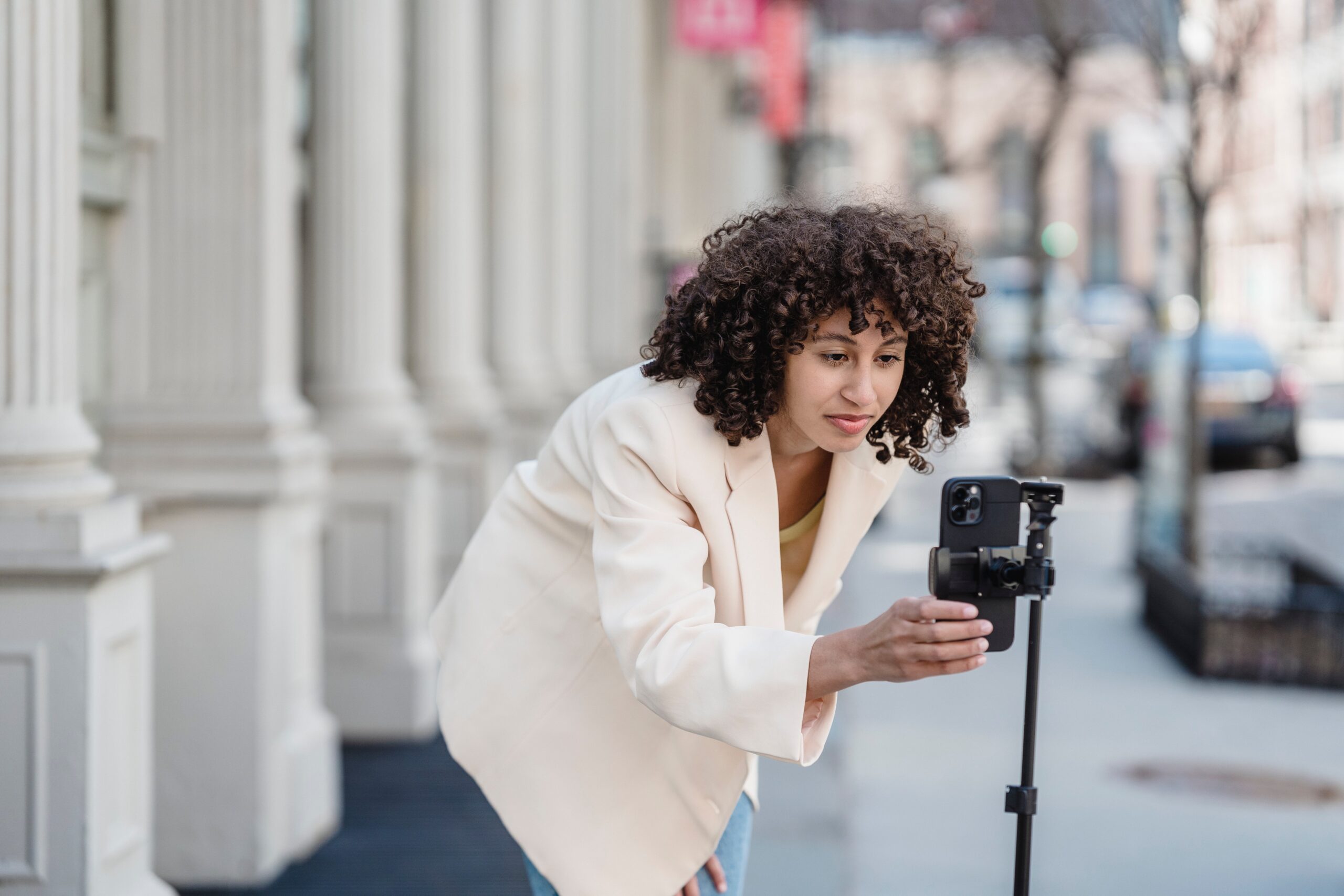 Content creator in a light-colored coat using a smartphone on a tripod to record a video on a city sidewalk, focused on adjusting the device.