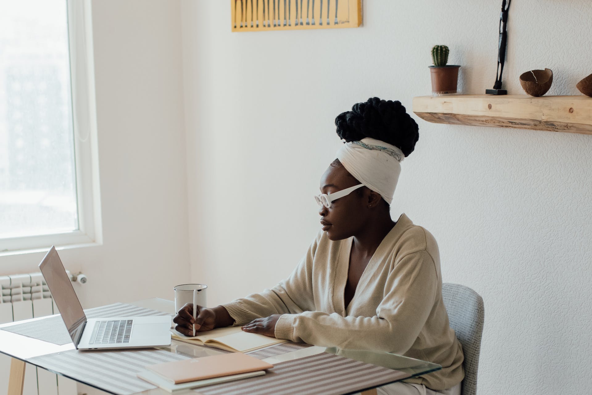 A focused woman wearing a headband sits at a minimalist desk with a laptop, notepad, and a cup, embracing authenticity as she works diligently in a bright, cozy workspace.