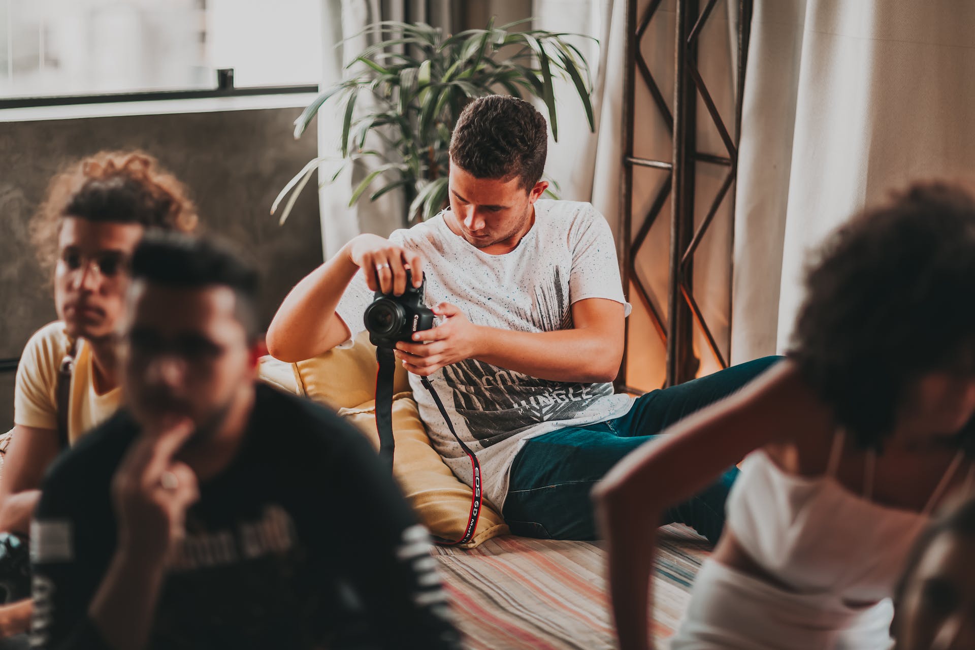 Group of young adults in a casual indoor setting, with one man intently focusing on a camera in his hands. He is seated on a couch, reviewing his camera settings or captured images, oblivious to his friends blurred in the background. This moment captures the essence of dedication and concentration, a visual metaphor for the concept 'Likes Don't Pay,' highlighting the importance of substance and skill over mere social media validation.