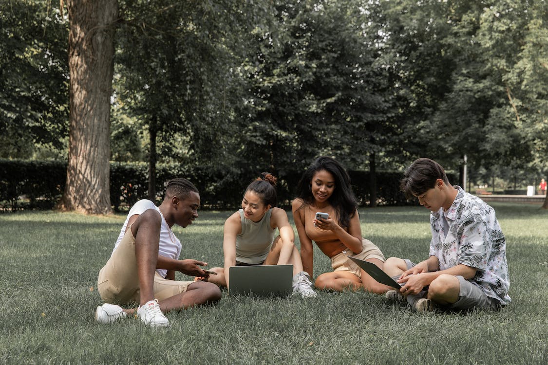 A group of four diverse young adults sitting on the grass in a park, collaboratively working on a laptop and smartphones. They appear to be thoughtfully discussing and rethinking their intent to enhance social media engagement, embodying the spirit of connectedness and strategic planning in a serene outdoor setting.