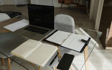 A modern workspace featuring an open laptop on a glass table, with a planner opened to a blank page alongside a pen, all ready for planning. A smartphone rests nearby, symbolizing the integration of digital and traditional planning. This serene setting invites contemplation on the question: What will you do to make this a successful year?