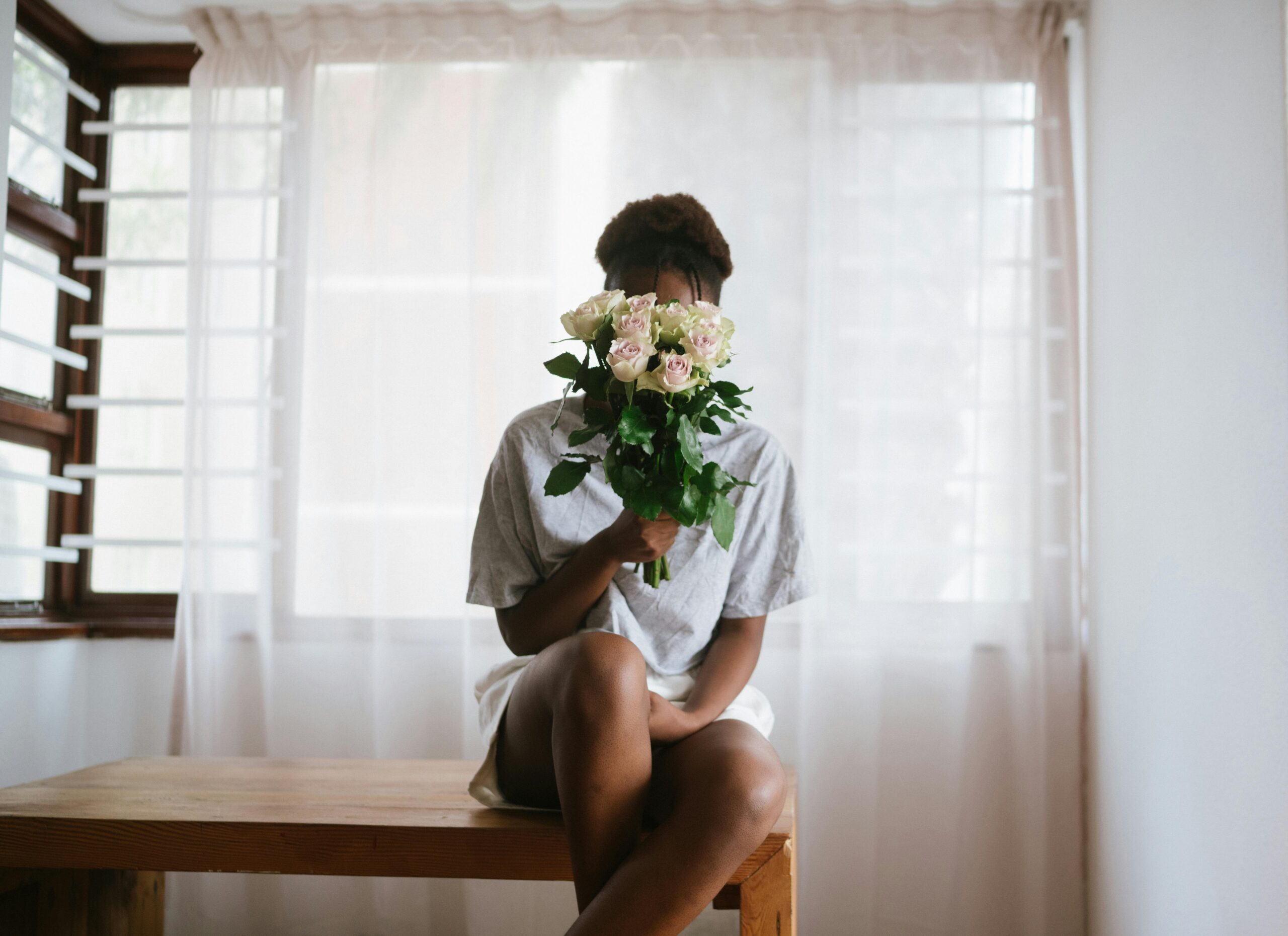 A person sitting serenely on a wooden stool, holding a bouquet of fresh, blooming flowers close to their face, symbolizing a moment of self-care and the practice of intentional love.